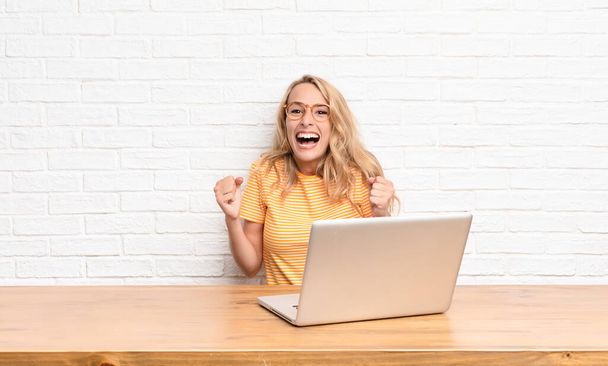 young blonde woman feeling shocked, excited and happy, laughing and celebrating success, saying wow! using a laptop - Fotoğraf, Görsel