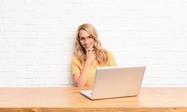young blonde woman smiling, enjoying life, feeling happy, friendly, satisfied and carefree with hand on chin using a laptop - Photo, Image