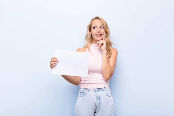 young blonde woman smiling happily and daydreaming or doubting, looking to the side holding a sheet of paper - Foto, afbeelding