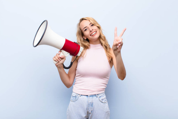 young blonde woman smiling and looking happy, carefree and positive, gesturing victory or peace with one hand with a megaphone - Fotoğraf, Görsel