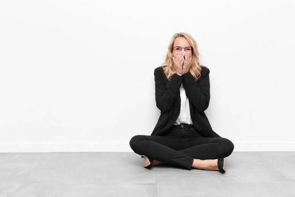 young blonde woman looking happy, cheerful, lucky and surprised covering mouth with both hands sitting on the floor - Photo, Image