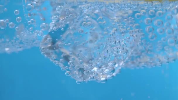 Extreme close up κομμάτια πάγου και φέτες λεμονιού σε μεταλλικό νερό σε μπλε φόντο. Φυσικό ανθρακούχο μεταλλικό νερό σε ένα ποτήρι. Φυσαλίδες αερίου στον πάγο. - Πλάνα, βίντεο