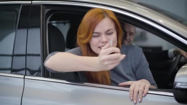 Portrait of beautiful redhead Caucasian woman holding car keys and making victory gesture. Joyful mother sitting in new automobile with husband and son. Auto industry, joy, happiness. - Séquence, vidéo