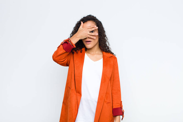young black woman looking shocked, scared or terrified, covering face with hand and peeking between fingers against white wall - Photo, Image