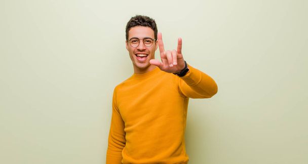 young arabian man feeling happy, fun, confident, positive and rebellious, making rock or heavy metal sign with hand against flat wall - Photo, image