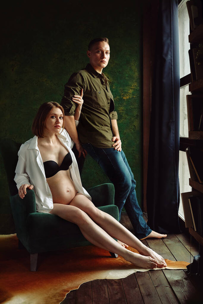 A pregnant couple is sitting on a chair by the window. Fashion Pregnancy Concept. - Фото, зображення