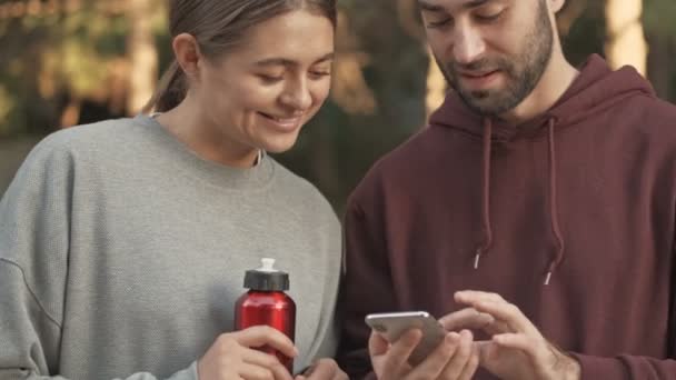 Close up άποψη της ευχαριστημένος νεαρό όμορφο ζευγάρι χρησιμοποιώντας smartphone και ανοίγοντας το δρόμο σε εξωτερικούς χώρους πάρκο - Πλάνα, βίντεο