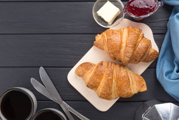 Fresh croissants on a black wooden table. Served with coffee, coffeepot, butter, jam, blue napkin. Fresh French Baked Croissants. Warm Fresh Buttery Rolls. Breakfast. Free space for text. View from above. Top view.  - Фото, изображение