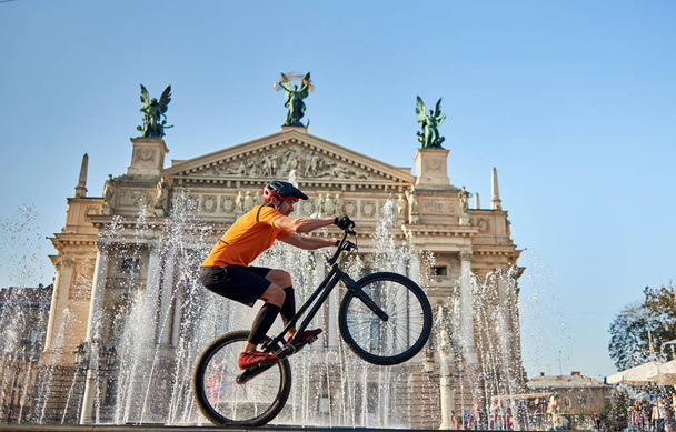Desportista extremo realizando passeio perigoso na roda traseira de bicicleta de montanha na frente da fonte e casa de ópera, vista lateral
 - Foto, Imagem