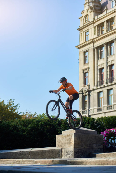 Egy bátor ember oldalnézete, aki biciklivel trükközik a belvárosban. Fiatal kerékpáros sisakban és narancssárga ingben lovagol az építészet hátterében. A szélsőséges fogalom. - Fotó, kép