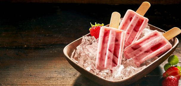 Refreshing frozen fresh strawberry popsicles displayed in a bowl of ice over a rustic wooden counter with dark background and copy space in panorama banner format - Photo, image