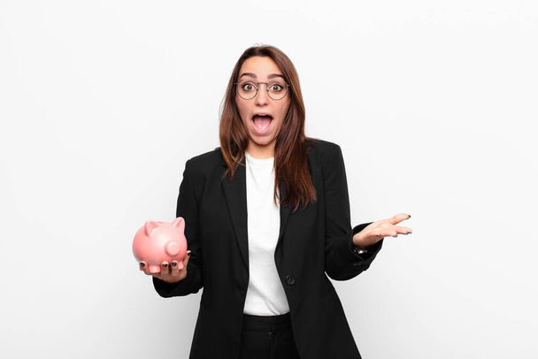 young businesswoman feeling extremely shocked and surprised, anxious and panicking, with a stressed and horrified look with a piggy bank - Photo, image