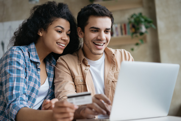 Portrait of beautiful multiracial couple shopping online, using laptop computer.  Happy friends ordering food on website. Smiling African America woman holding credit card, looking at digital screen  - Fotoğraf, Görsel