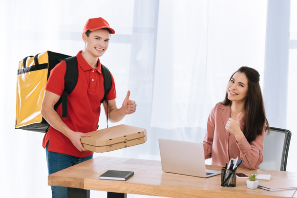 Delivery man with pizza boxes showing thumbs up gesture near businesswoman at table - Foto, Imagen