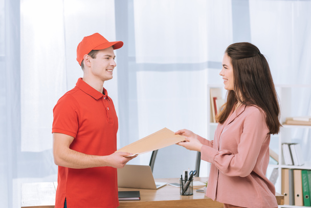 Side view of delivery man giving envelope to smiling businesswoman in office  - Φωτογραφία, εικόνα