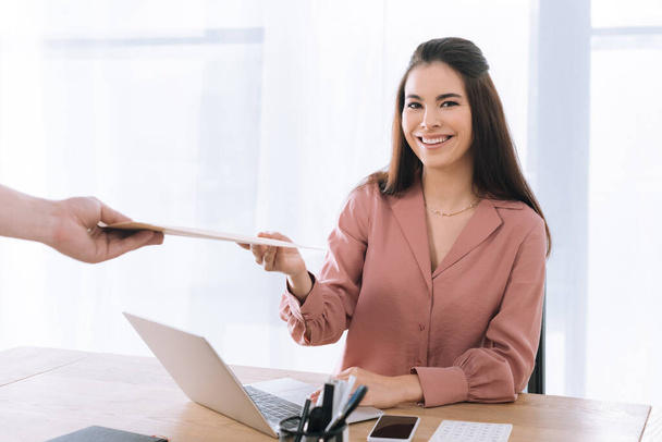Attractive businesswoman smiling at camera near courier with envelope in office - Zdjęcie, obraz