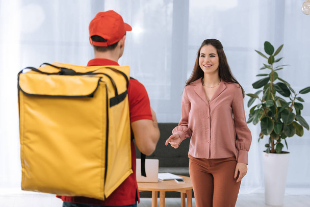 Selective focus of businesswoman smiling at courier with thermo backpack in office   - Photo, Image