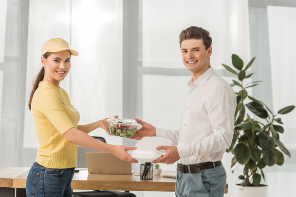 Side view of courier giving food containers to businessman and smiling at camera in office - Фото, изображение