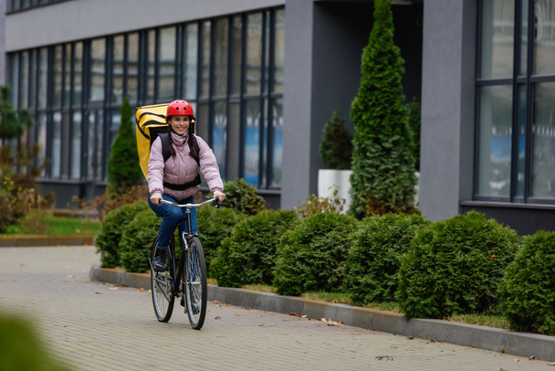 Enfoque selectivo de mensajero sonriente con mochila termo a caballo en bicicleta en la calle urbana
  - Foto, Imagen