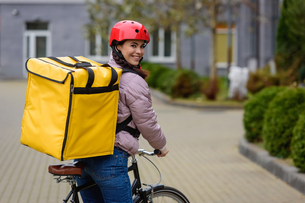 Vista lateral del atractivo mensajero con mochila térmica y bicicleta sonriendo a la cámara en la calle
 - Foto, Imagen
