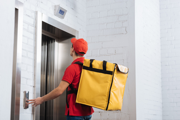 Side view of delivery man with thermo backpack pressing elevator button  - Foto, Imagen