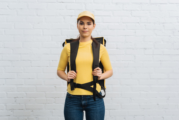 Attractive courier in yellow uniform with thermal backpack looking at camera near brick wall - 写真・画像