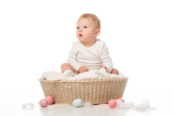 Child with open mouth sitting on blanket in basket with Easter eggs around on white background - Photo, Image