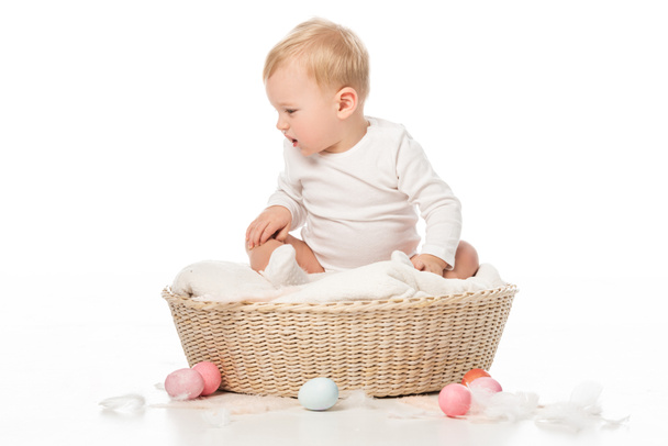 Dítě dívá dolů a sedí v košíku s velikonočními vejci kolem na bílém pozadí - Fotografie, Obrázek