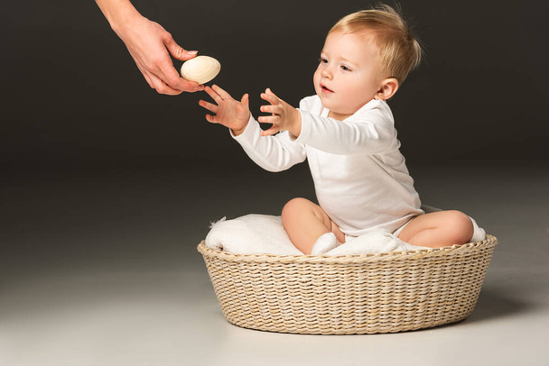 Vista cortada de menino bonito tomando ovo de Páscoa da mão da mulher, sentado em cobertor na cesta no fundo preto
 - Foto, Imagem