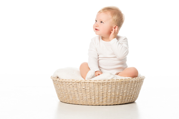 Kind berührt Ohr und schaut weg, sitzt auf Decke in Korb auf weißem Hintergrund - Foto, Bild