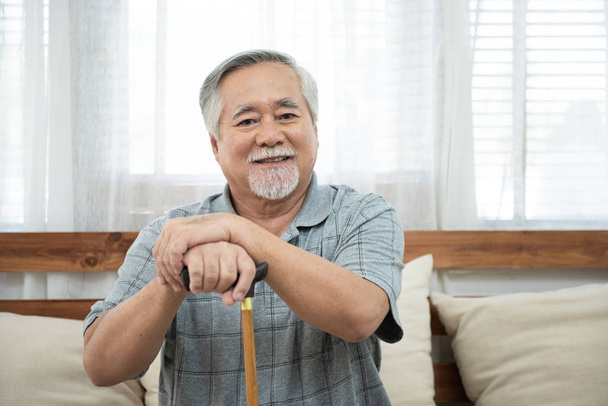 Portrait of senior old elderly asian man sit on coach hand hold help walking stick sit on sofa in house look at camera with happiness and healthy lifestyle. - Photo, Image