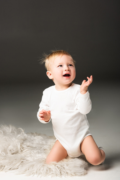 niedliches glückliches Kind, das mit offenem Mund nach oben schaut, auf Fell auf schwarzem Hintergrund sitzend - Foto, Bild