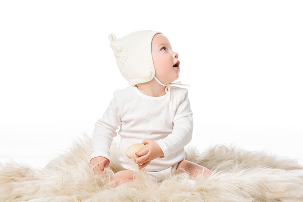 Kid wearing baby bonnet, holding easter egg, looking away with open mouth on fur isolated on white  - 写真・画像