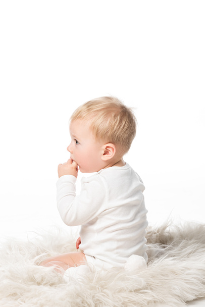 Πλευρική άποψη του χαριτωμένο παιδί με το δάχτυλο στο στόμα, κάθεται σε γούνα που απομονώνονται σε λευκό  - Φωτογραφία, εικόνα