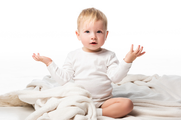 niedliches Kind, das die Hände zur Seite spreizt und auf einer Decke auf weißem Hintergrund sitzt - Foto, Bild