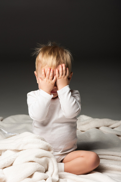Cute child covering face with hands and sitting on blanket on black background - Foto, afbeelding