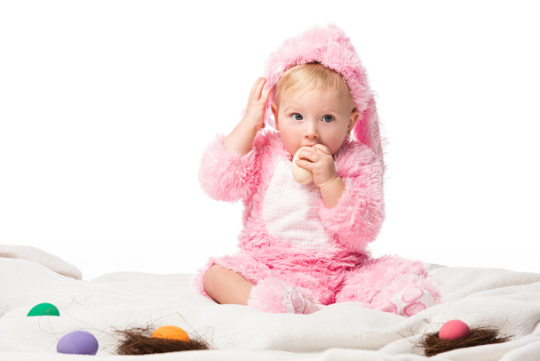 Child wearing rabbit costume, putting easter egg in mouth, touching head on blanket isolated on white  - Photo, Image