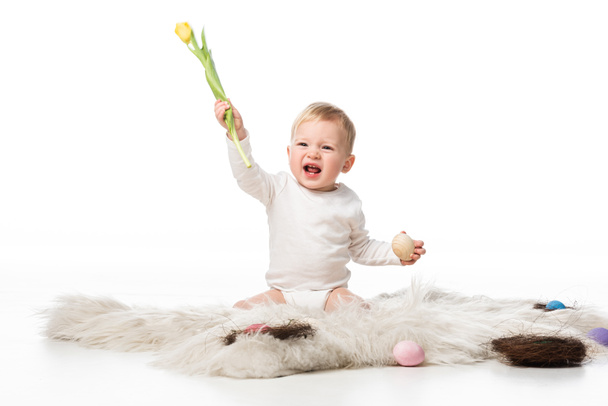 Kind mit offenem Mund hält Tulpe und Osterei, sitzt auf Fell auf weißem Hintergrund - Foto, Bild