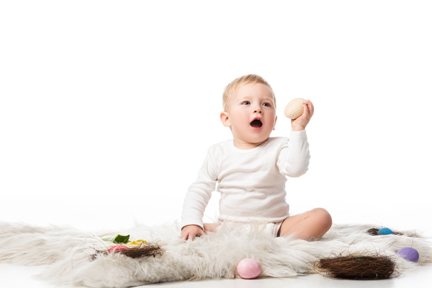 Ağzı açık bir çocuk, elinde paskalya yumurtası, beyaz arka planda kürke oturuyor. - Fotoğraf, Görsel