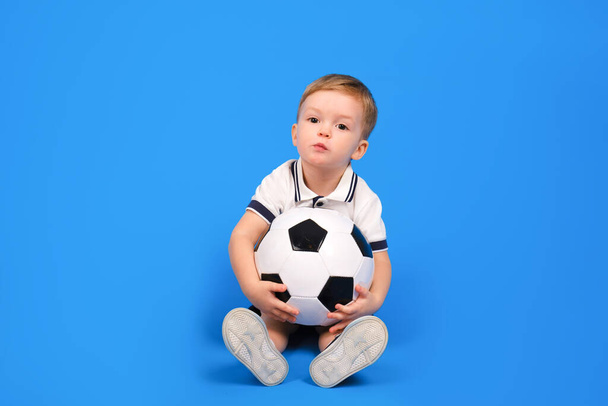 Jongen in een voetbal uniform zit op een blauwe achtergrond met een voetbal en kijkt vooruit. - Foto, afbeelding