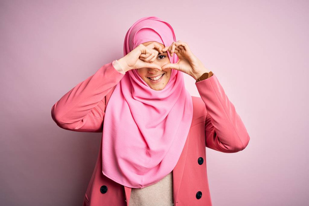 Nuori kaunis tyttö yllään muslimi hijab seisoo yli eristetty vaaleanpunainen tausta tekee sydämen muodon käsin ja sormin hymyillen läpi merkki
 - Valokuva, kuva