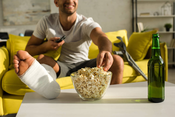 Cropped view of smiling man with broken leg taking popcorn near beer bottle white watching tv at home - Photo, Image