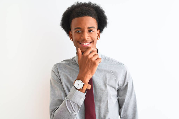 Joven hombre de negocios afroamericano con corbata de pie sobre un fondo blanco aislado mirando confiado a la cámara con sonrisa con los brazos cruzados y la mano levantada en la barbilla. Pensando positivamente
. - Foto, imagen