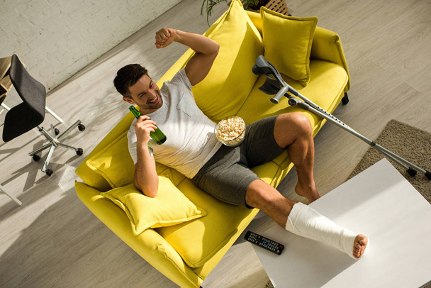 Vysoký úhel pohledu na veselý muž se zlomenou nohou sledování televize s popcornem a pivem na gauči - Fotografie, Obrázek