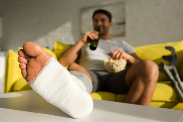 Enfoque selectivo del hombre con la pierna rota bebiendo cerveza y comiendo palomitas de maíz en el sofá en la sala de estar
 - Foto, Imagen
