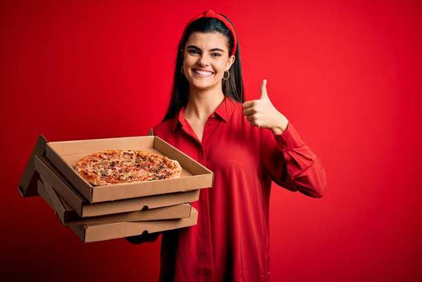 Jeune belle femme brune tenant des boîtes de livraison avec pizza italienne sur fond rouge heureux avec un grand sourire faisant signe ok, pouce levé avec les doigts, excellent signe
 - Photo, image