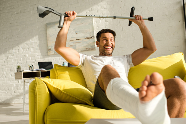 Селективный фокус позитивного человека со сломанной ногой, поднимающей костыль, сидя дома на диване
 - Фото, изображение