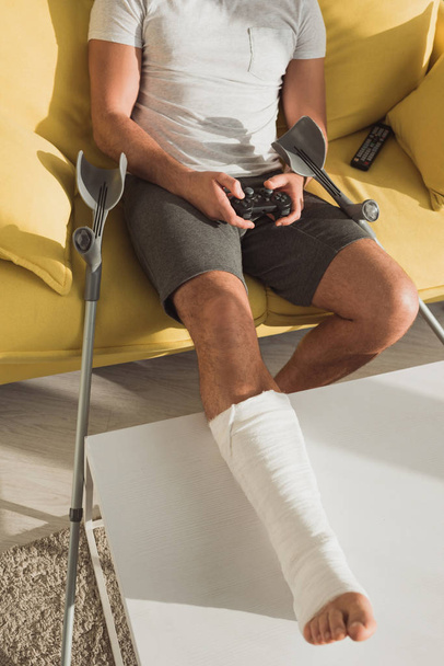 KYIV, UKRAINE - JANUARY 21, 2020: Cropped view of man with broken leg playing video game near crutches and remote controller on couch - Zdjęcie, obraz