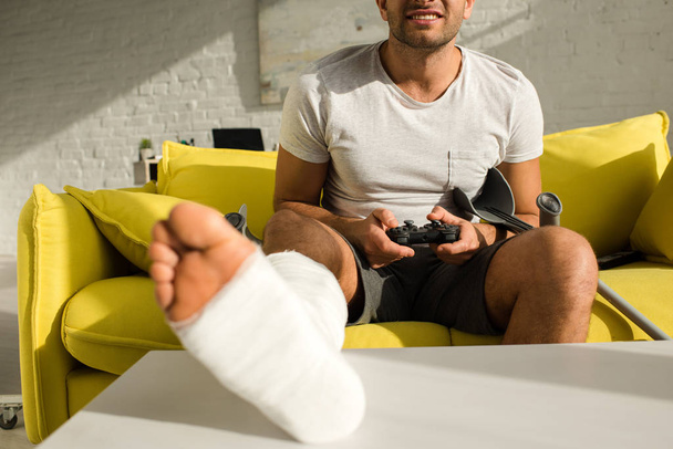 KYIV, UKRAINE - JANUARY 21, 2020: Selective focus of smiling man with leg in plaster bandage on coffee table playing video game at home - Fotografie, Obrázek