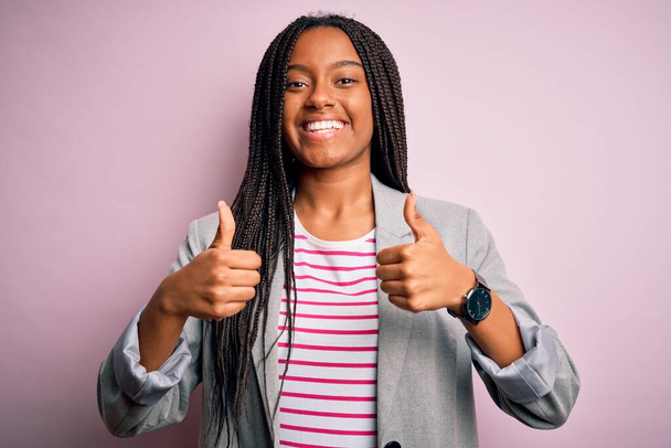 Jovem mulher de negócios afro-americana em pé sobre rosa isolado sinal de sucesso fundo fazendo gesto positivo com a mão, polegares para cima sorrindo e feliz. Expressão alegre e gesto vencedor
. - Foto, Imagem